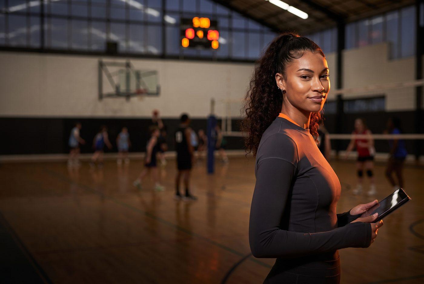 League organizer holding a tablet courtside while a volleyball league plays behind her